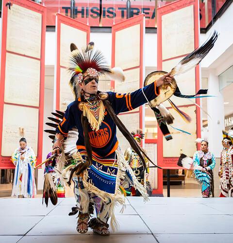 Indigenous dancer in feathered regalia performing a powwow demo in the mall