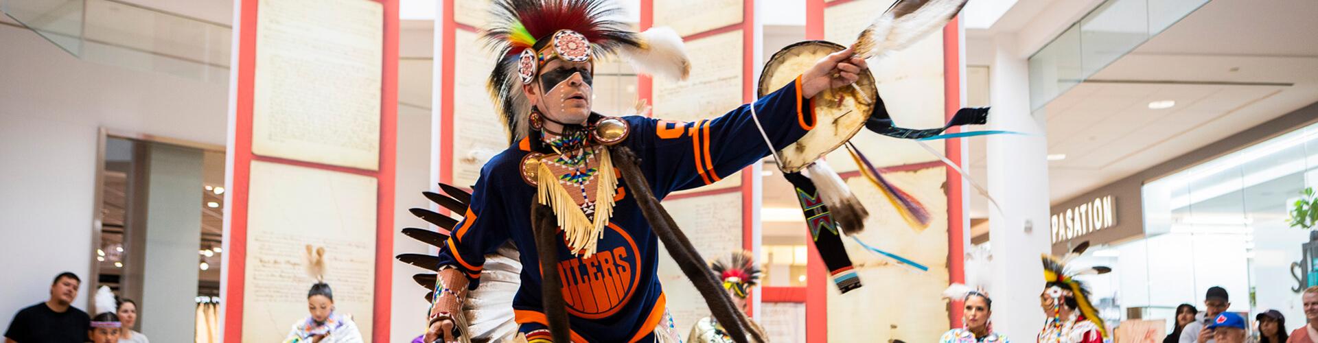 Indigenous dancer in feathered regalia performing a powwow demo in the mall