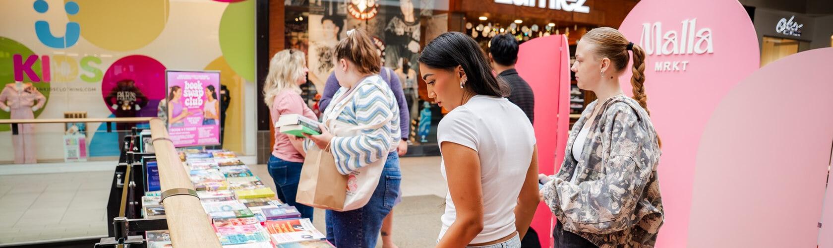 Shoppers browsing a book swap table at a pink Malla Mrkt mall pop-up