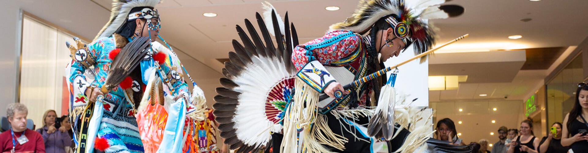Two Indigenous dancers in feathered regalia performing before a mall crowd