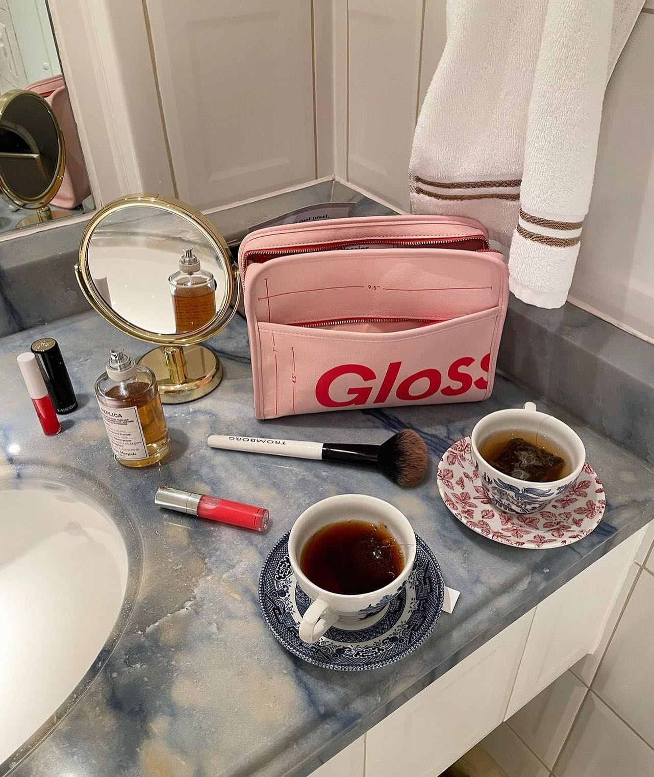 A Glossier make up bag sits on a bathroom countertop alongside two cups of tea and other Glossier products.