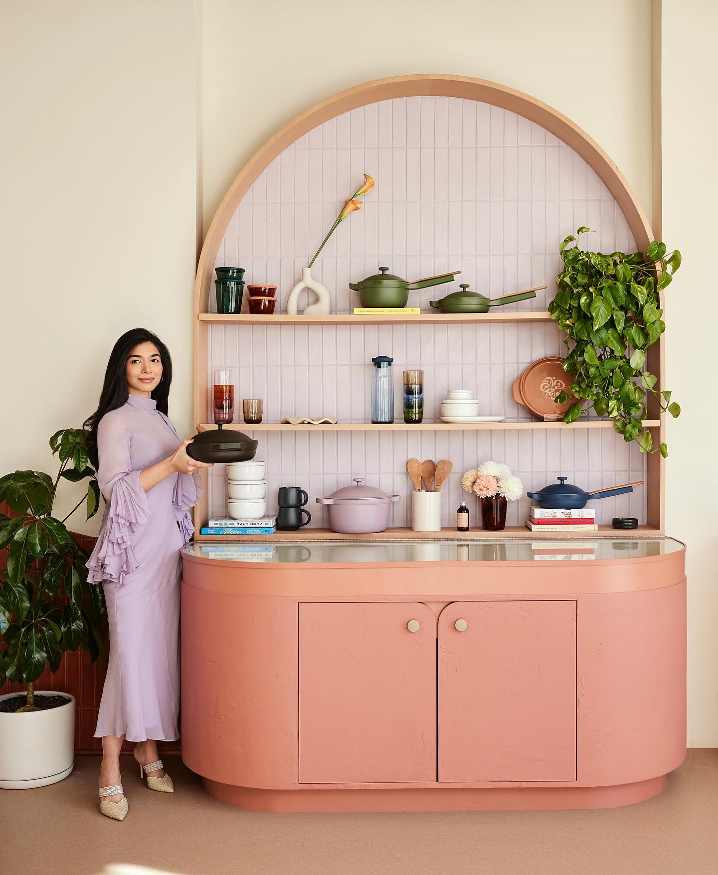 A woman is holding an Our Place pot. She is to the left of a retro-inspired display of other Our Place products.