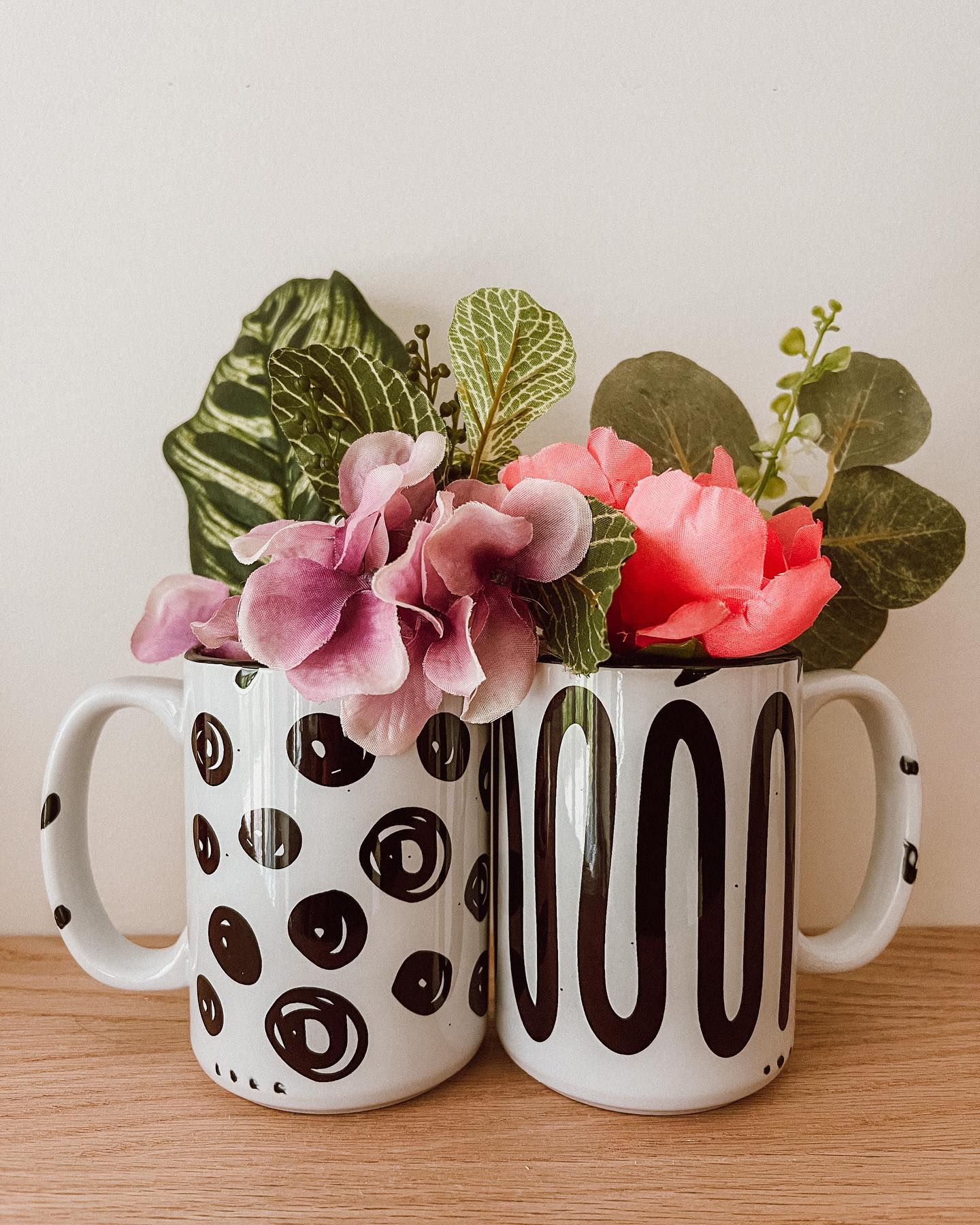 One spotted mug and one abstract line mug are side by side and filled with pink flowers.