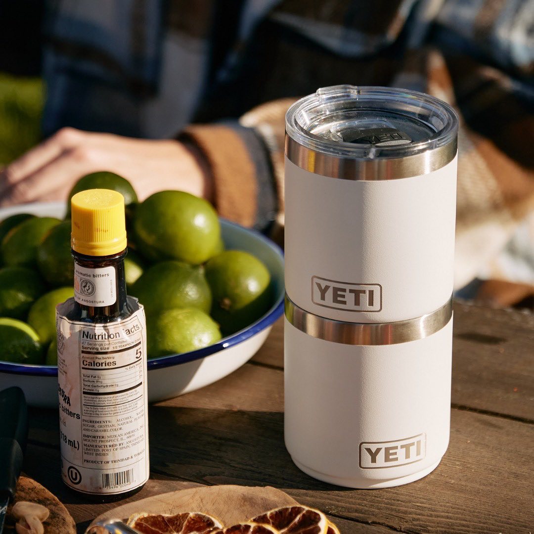 close up image of two stacked white YETI tumblers on a wooden table outdoors. The arm of a person wearing a plaid jacket can be seen in the background, and a small plate of limes is midground. Beside the yeti tumblers is a bottle of bitters with a yellow lid.