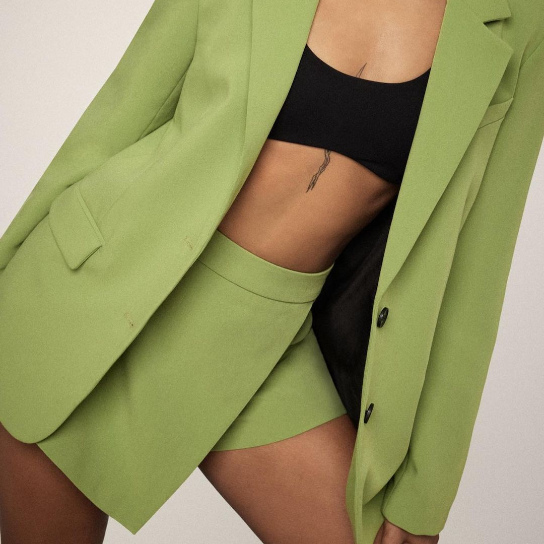 close-up image of a model wearing a matching green suit set consisting of a blazer and an asymmetrical skort. she wears a black bandeau bra underneath the blazer