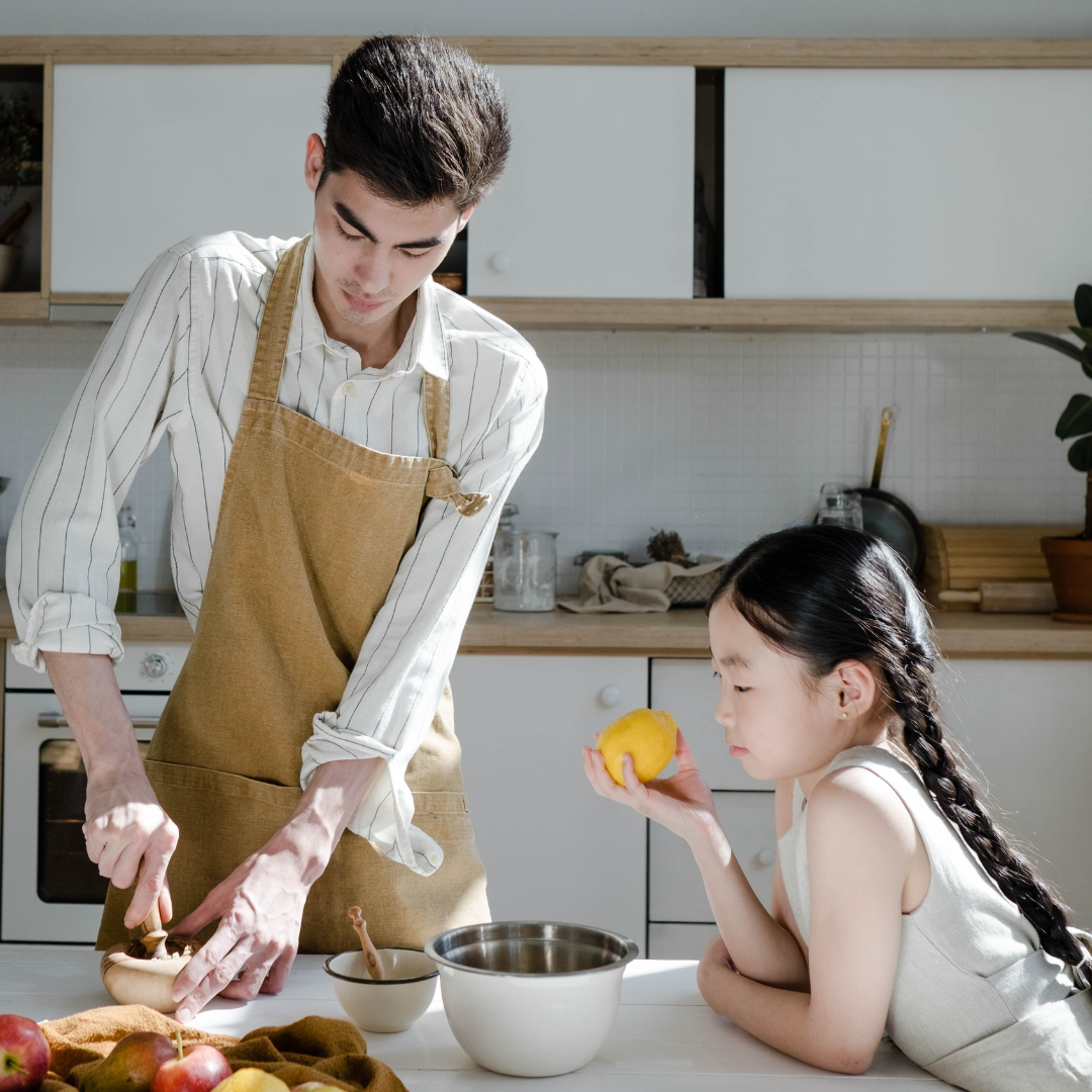 image of a man and a girl in the kitchen. the man is preparing food wearing a brown apron and the girl watches while holding a lemon