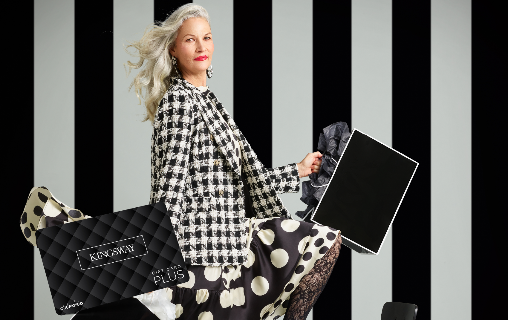 promotional image for a Kingsway gift card. It shows a woman wearing a black and white houndstooth blazer and black and white polka dot skirt holding black and white shopping bags. A Kingsway gift card is also in the image