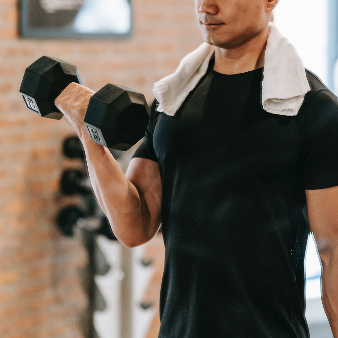 close-up image of a man in a gym lifting a dumbbell. he's wearing a black tshirt and a white towel around his neck