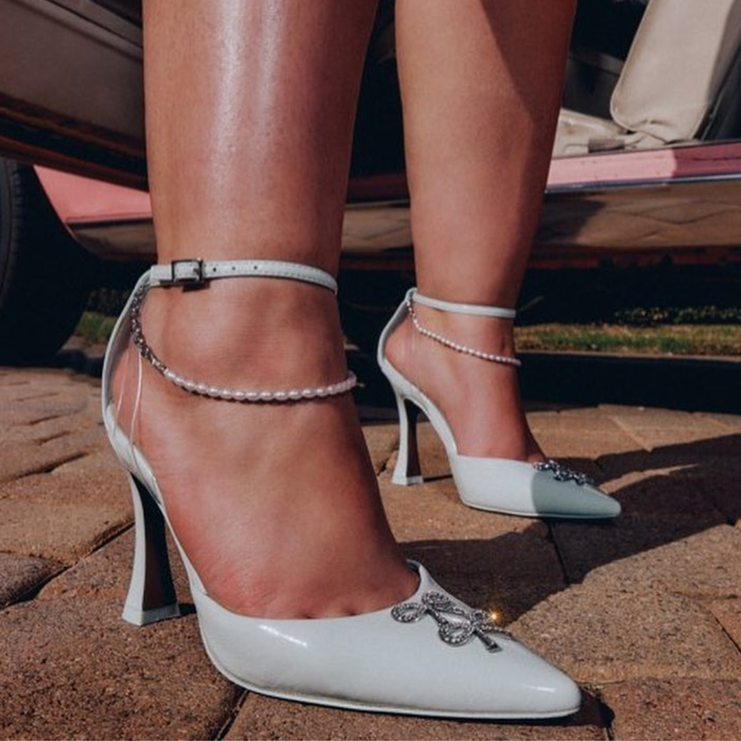 close-up image of a model earing pointy toed white shoes with two sparkly bows and a pearl strap detail