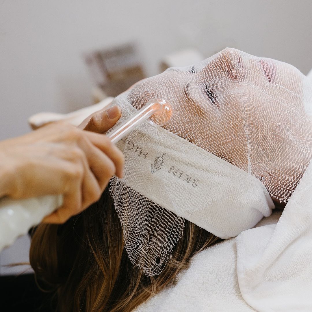 close up image of a woman getting a facial. she wears a white terry cloth headband and has gauze over her face while a technician's hand holding a wand can be seen hovering above her forehead