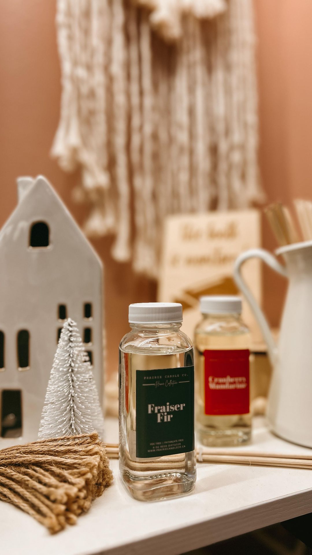 Holiday display at UNITE. In the foreground is a bottle of Fraiser Fir reed diffuser. In the background there is a white fir tree and a ceramic model of a white home.