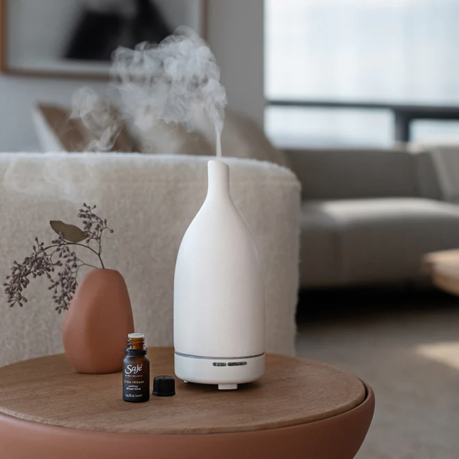 lifestyle image of a white saje diffuser with a diffuser blend and a small plant on a coffee table. there is a couch in the background
