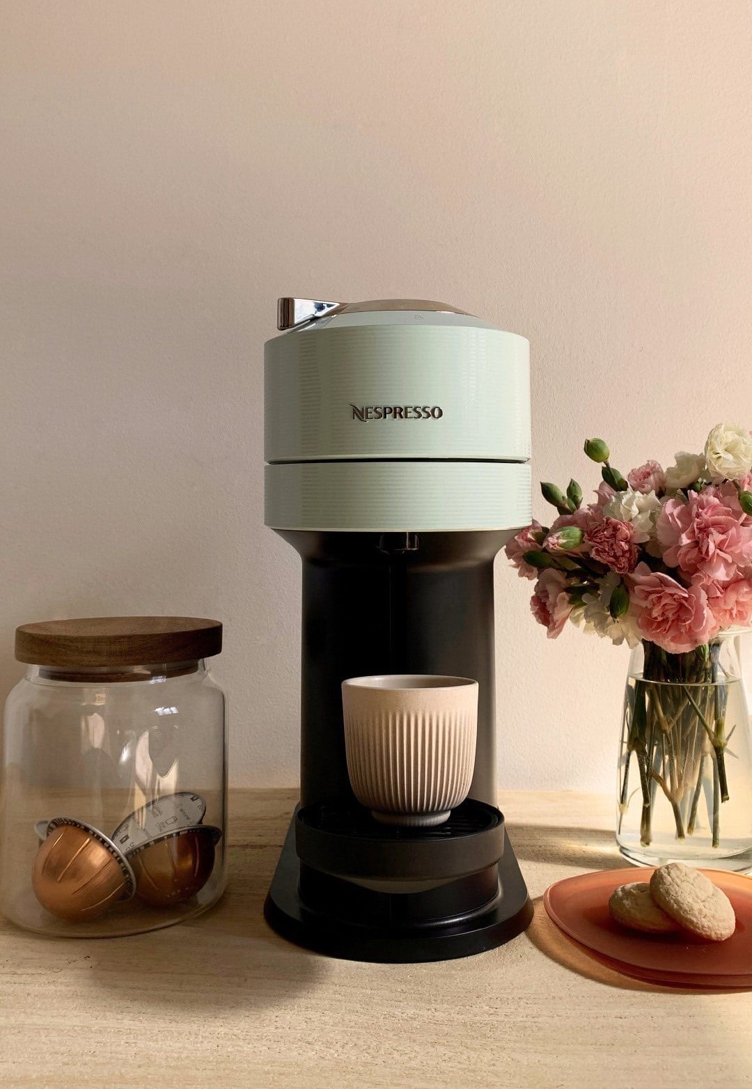 lifestyle image of a nespresso virtuo machine. to its right is a jar with nespresso pods and to the right there are pink and white florals and a plate with two cookies