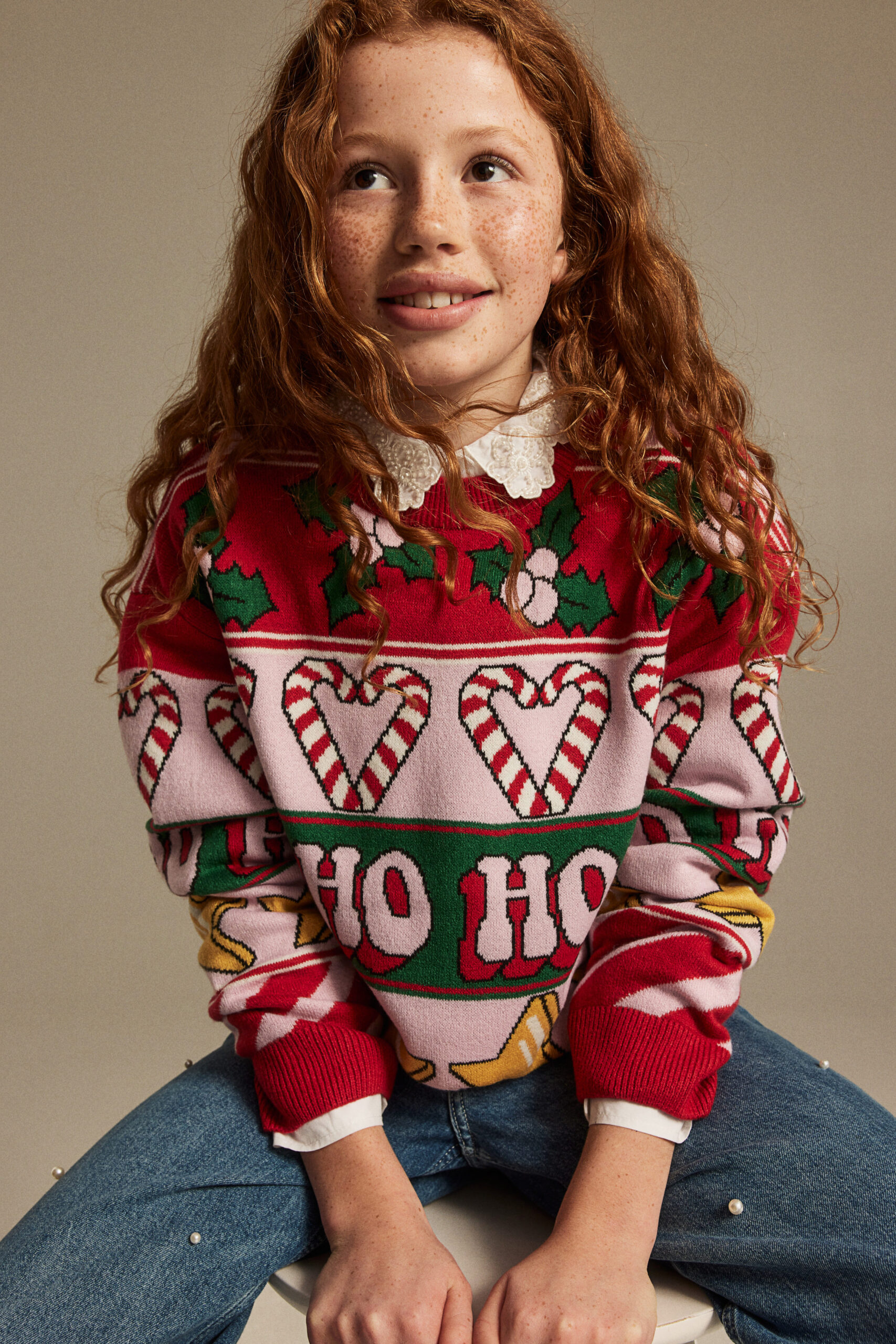 girl wearing jacquard-knit sweater in light pink/red/green with patterned mistletoe, hearts, stars, and "ho ho ho"