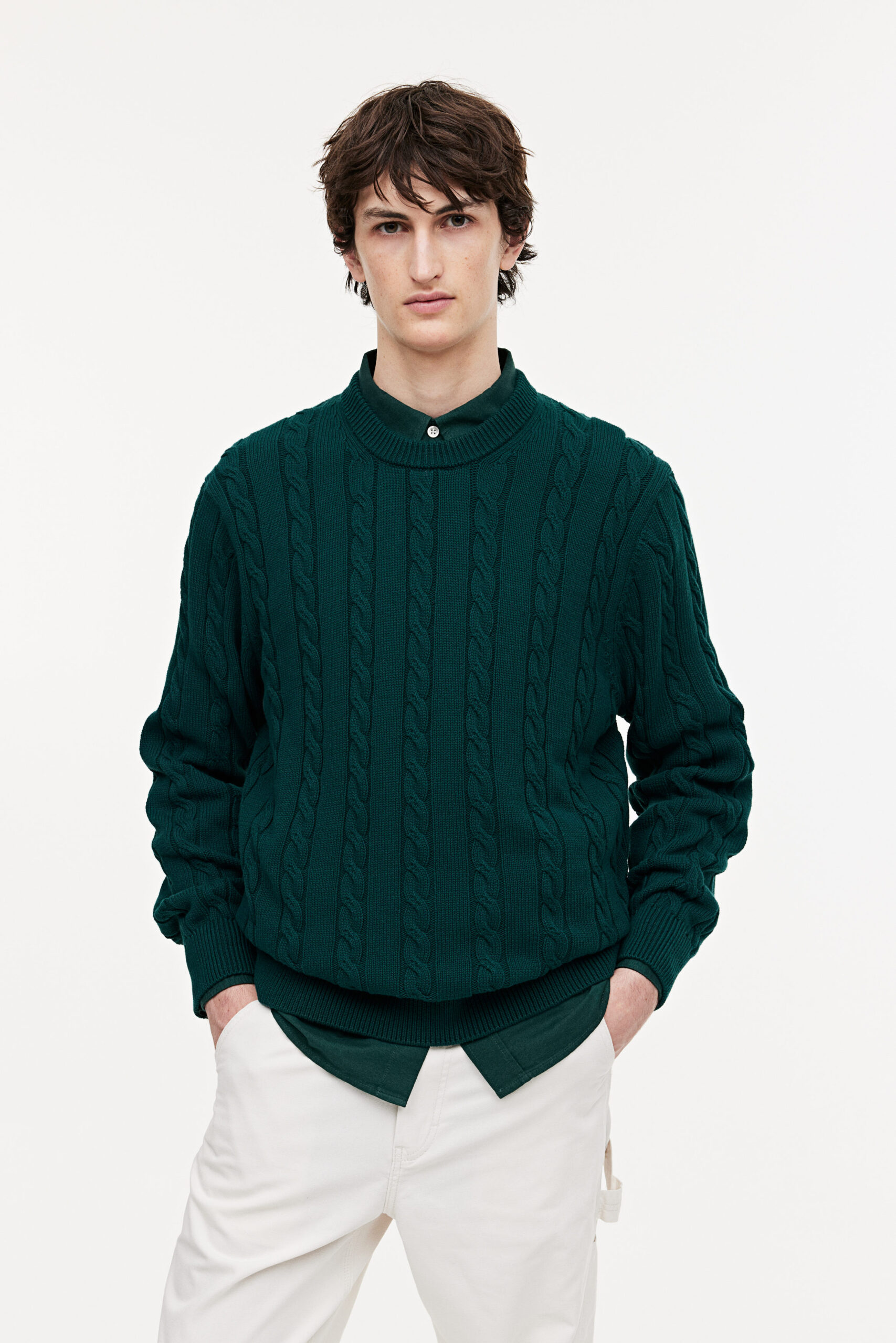 man wearing green knit sweater over a button up shirt with white pants