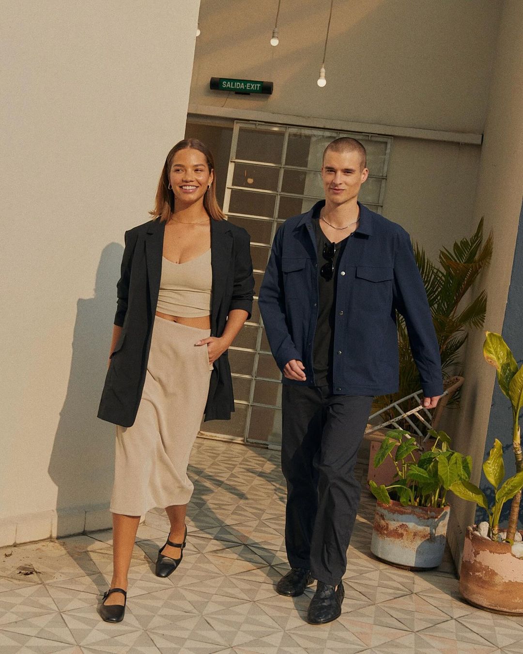 woman wearing tan coloured crop top and long skirt with blue blazer, and man wearing black shirt and pants with blue overshirt