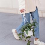 model wearing beige blazer, blue jeans and white sneakers, carrying potted plant while walking