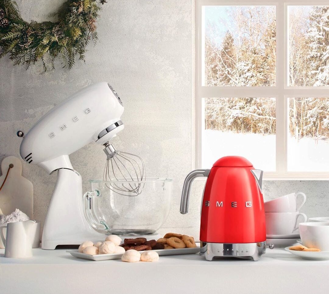 White stand mixer and red kettle on kitchen table, surrounded by food items